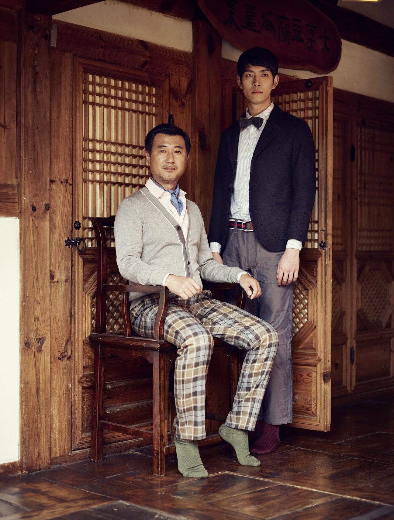 Two men in traditional Korean interior: seated man in cardigan and plaid trousers, standing man in dark jacket and bow tie.