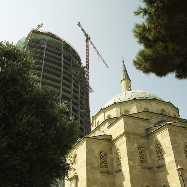 A traditional Islamic mosque with cream-colored stone facade stands beside a modern glass and steel high-rise building under construction with cranes.