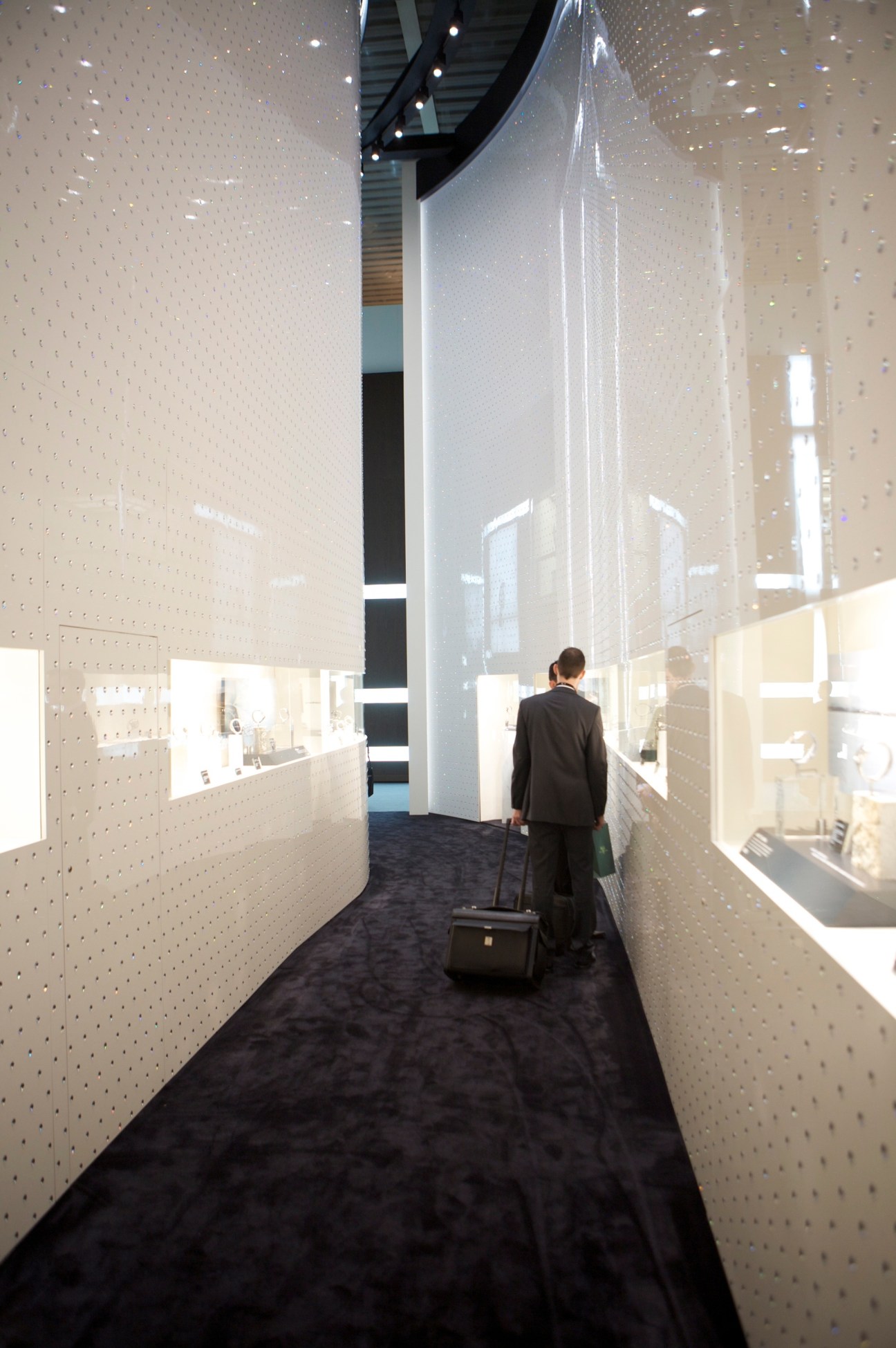 A man with luggage walks through a brightly lit corridor with illuminated display cases on either side at the Seiko stand.