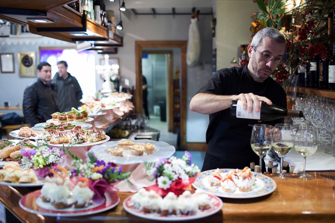 Esteban Ortega pours wine at Bergara bar with pintxos displayed on counter.