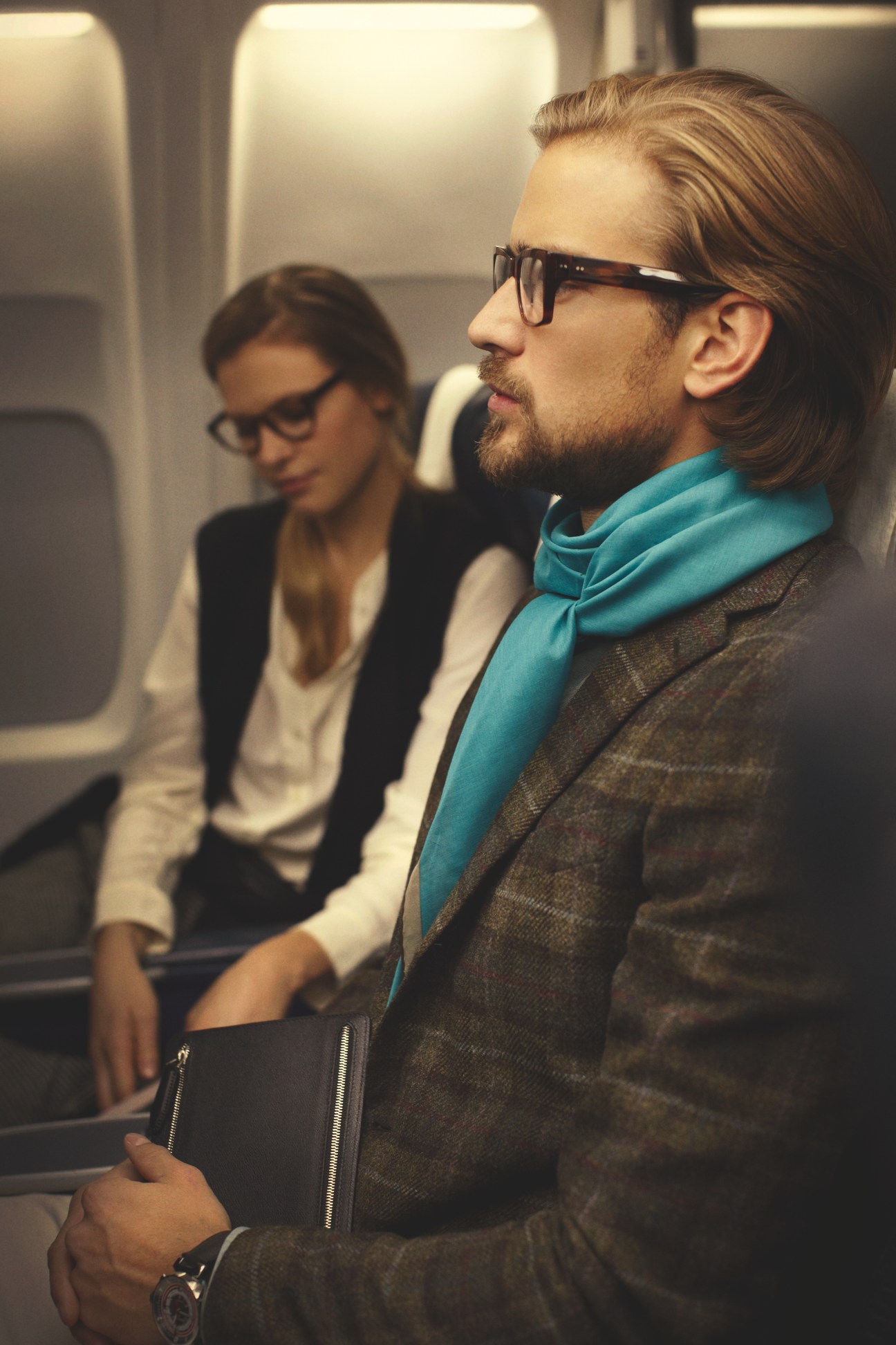 Man on airplane wearing Claire Goldsmith glasses, Hermès scarf, Caruso jacket and Smythson passport case.
