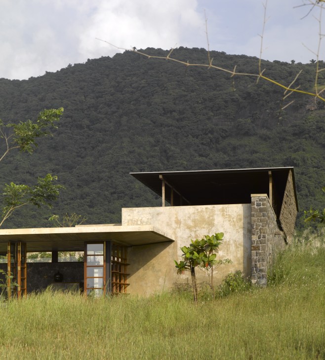 Modern house with ochre-colored walls and black basalt stone nestled in green grassland with forested hills beyond.
