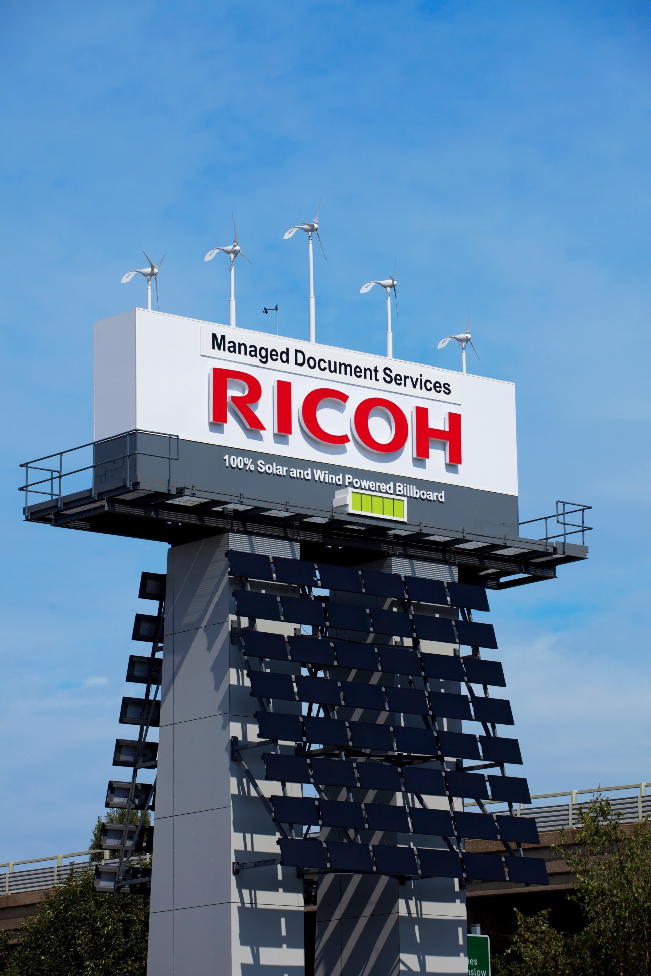 Solar and wind-powered eco-billboard displaying Ricoh branding against a blue sky.