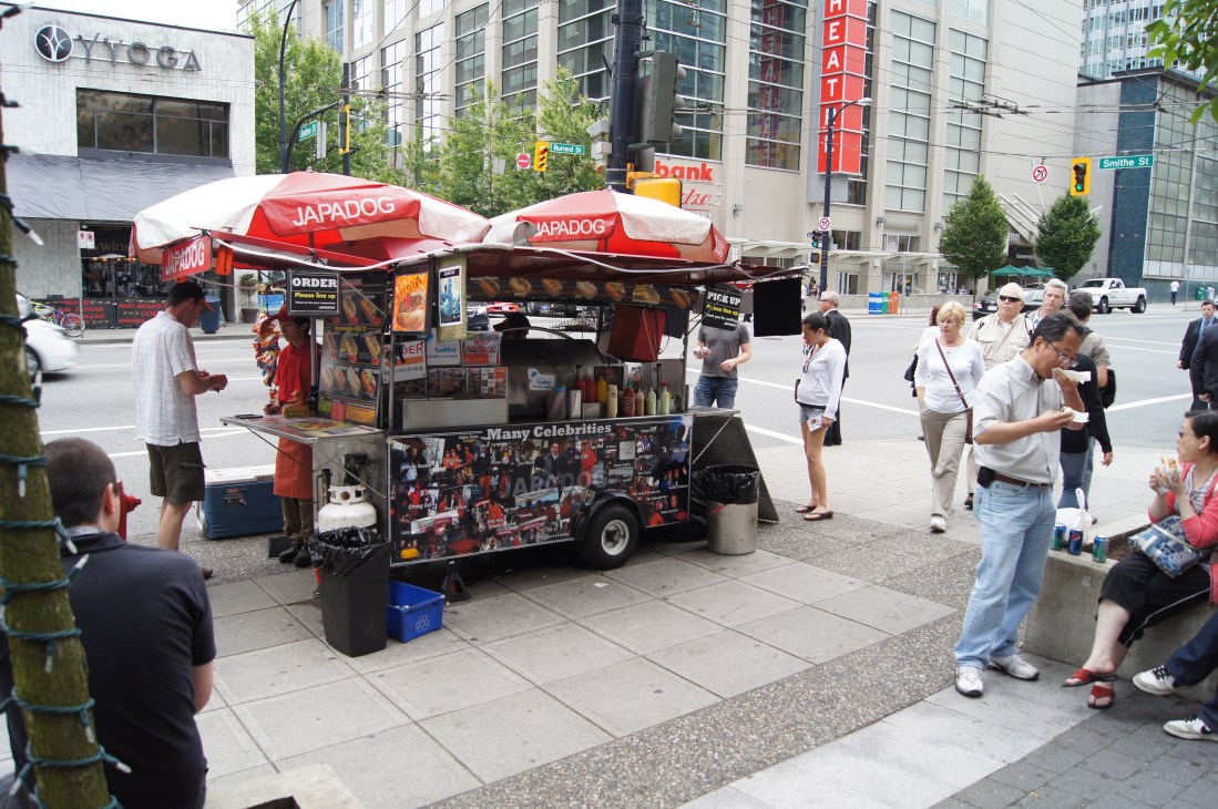 Japa Dog street food vendor cart with red umbrellas on a busy Vancouver street with pedestrians and urban buildings.