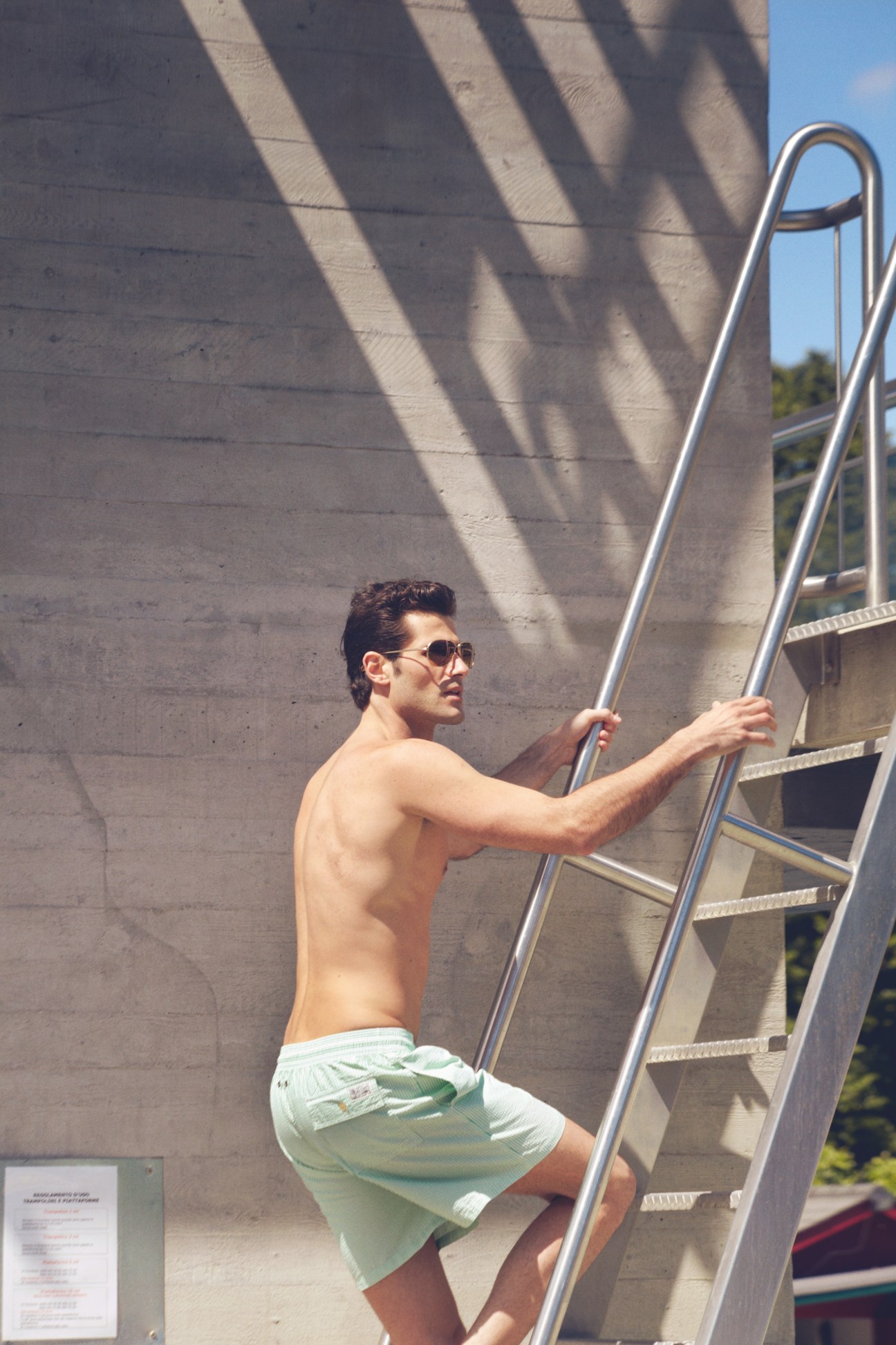 Man in sunglasses and swim trunks climbing a metal ladder against a concrete wall at a lido.