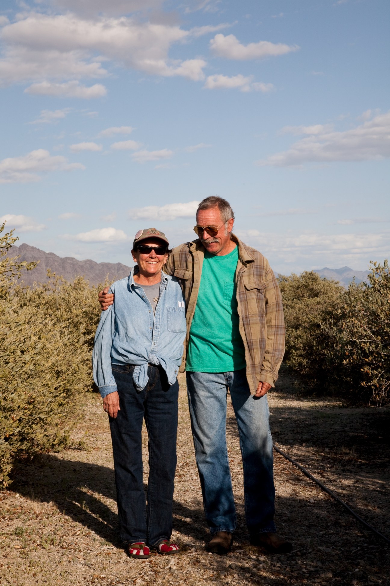 Larry and Donna Charpied stand together on a desert path surrounded by scrub vegetation with mountains in the background.