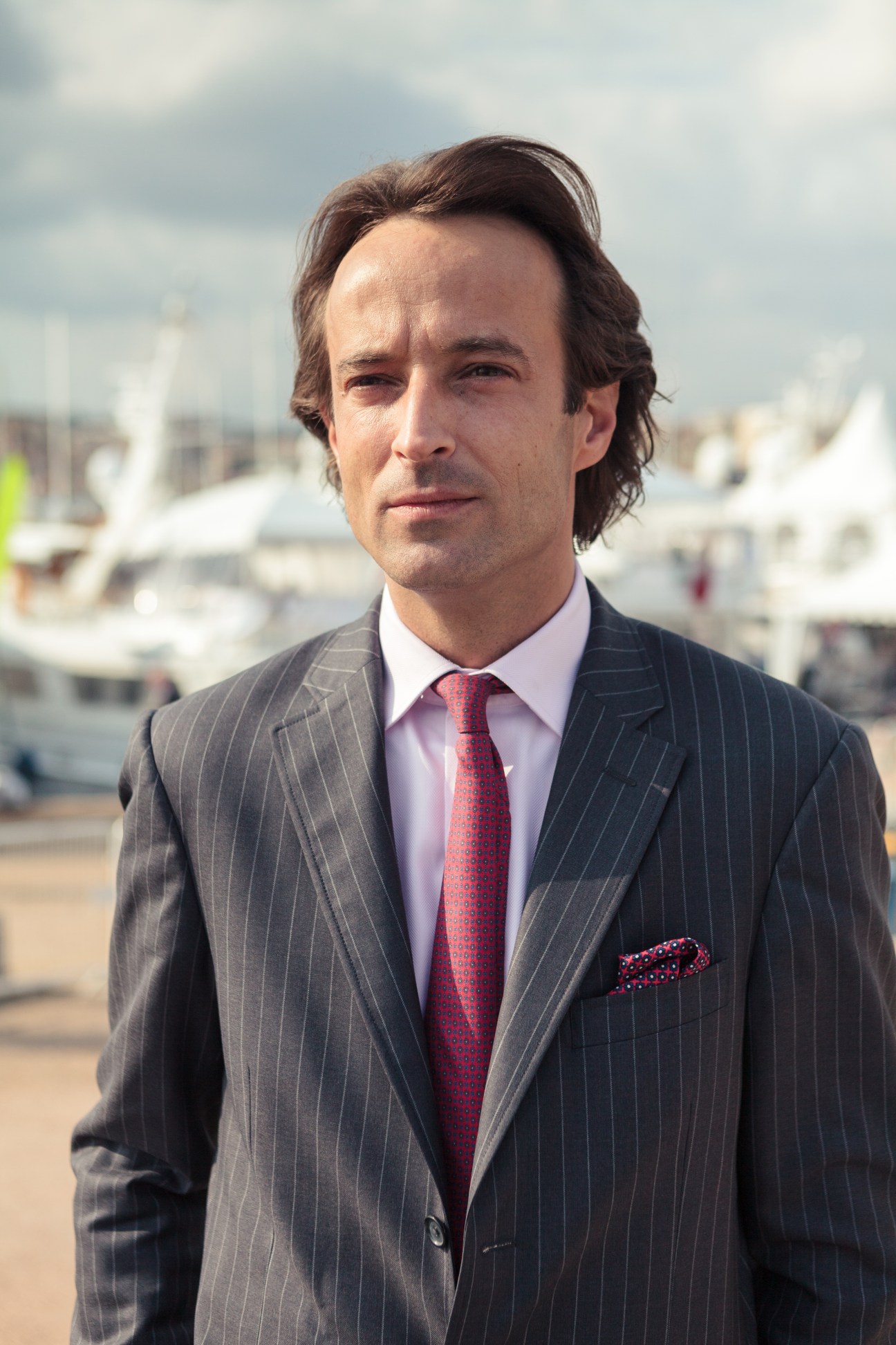Man in pinstriped suit and red tie standing in front of Cannes marina with yachts.