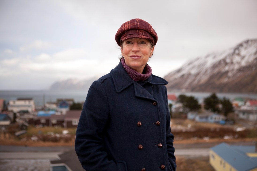 Mayor Shirley Marquardt stands outdoors wearing a burgundy cap and navy coat with Unalaska's harbor and mountains visible behind her.
