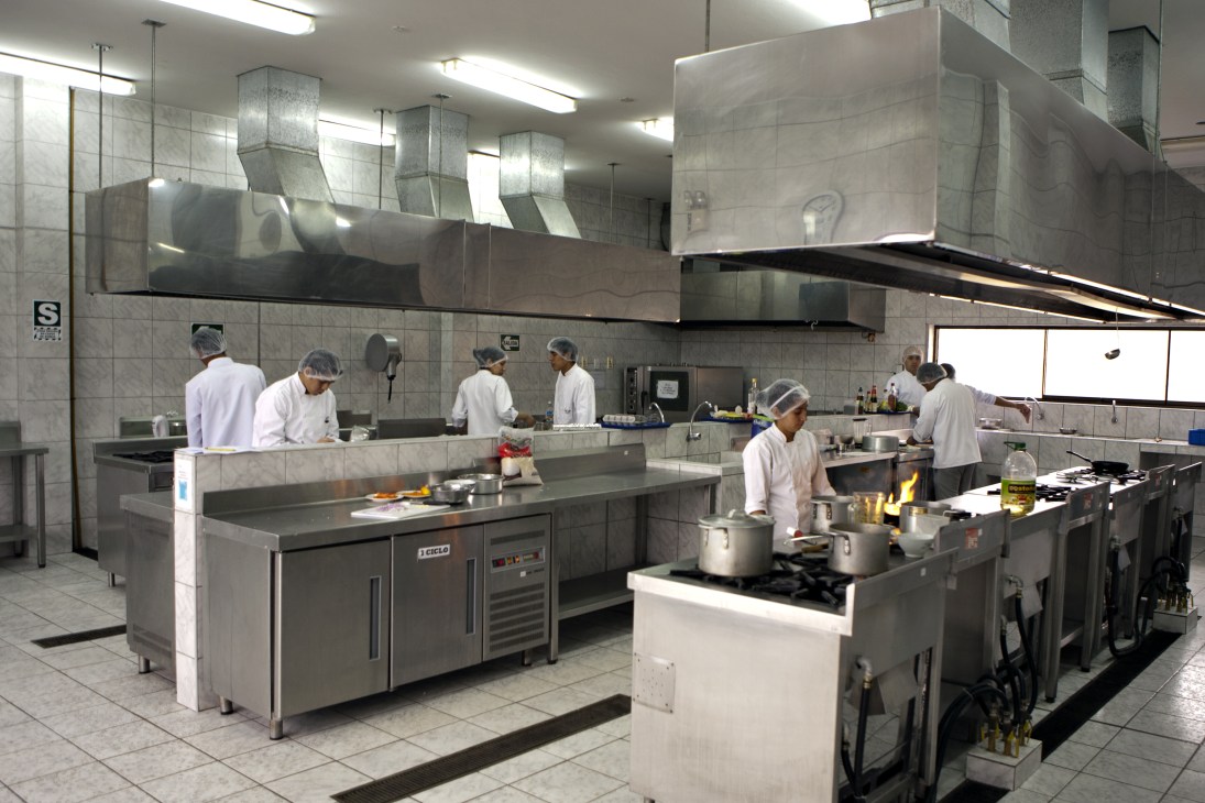 Students in white chef's uniforms work at multiple stainless steel cooking stations in a professional kitchen.