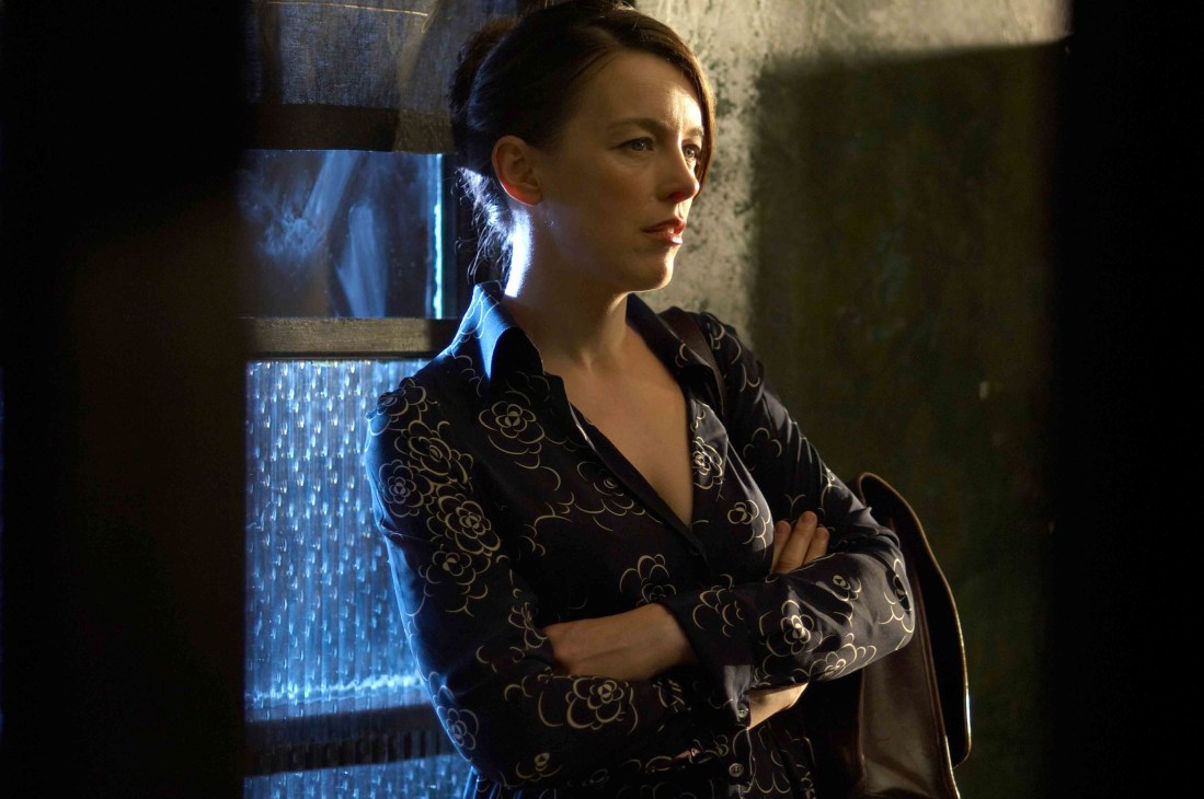 Woman in patterned black jacket standing against industrial wall with blue lighting.