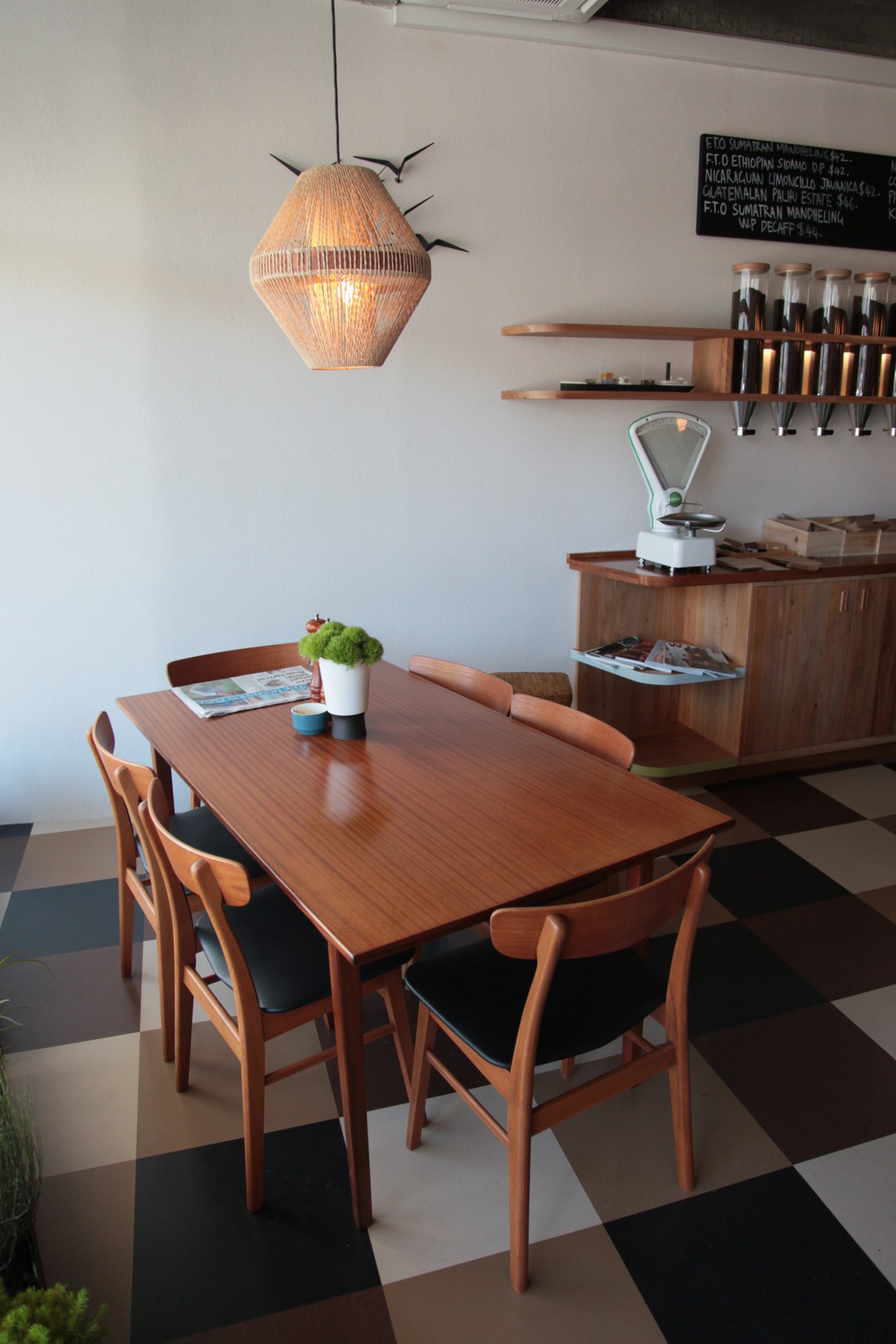 Interior of Customs coffee shop in Wellington featuring wooden dining table, mid-century chairs, woven pendant lamp and coffee equipment on shelves.