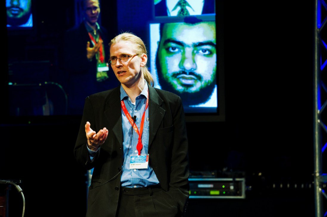 Mikko Hyppönen of F-Secure speaking at a conference with digital screens displaying faces in the background.