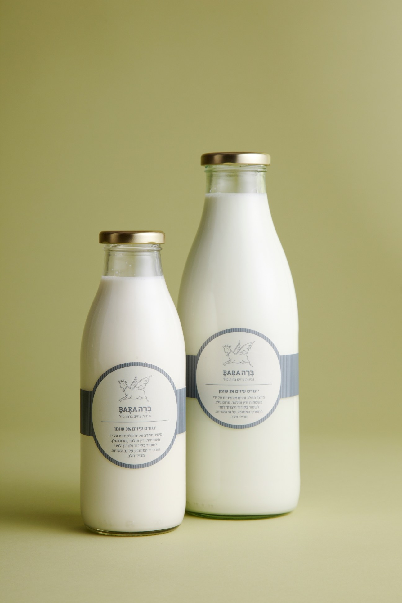 Two glass milk bottles with gold caps and circular labels featuring a flying goat illustration on a pale yellow background.