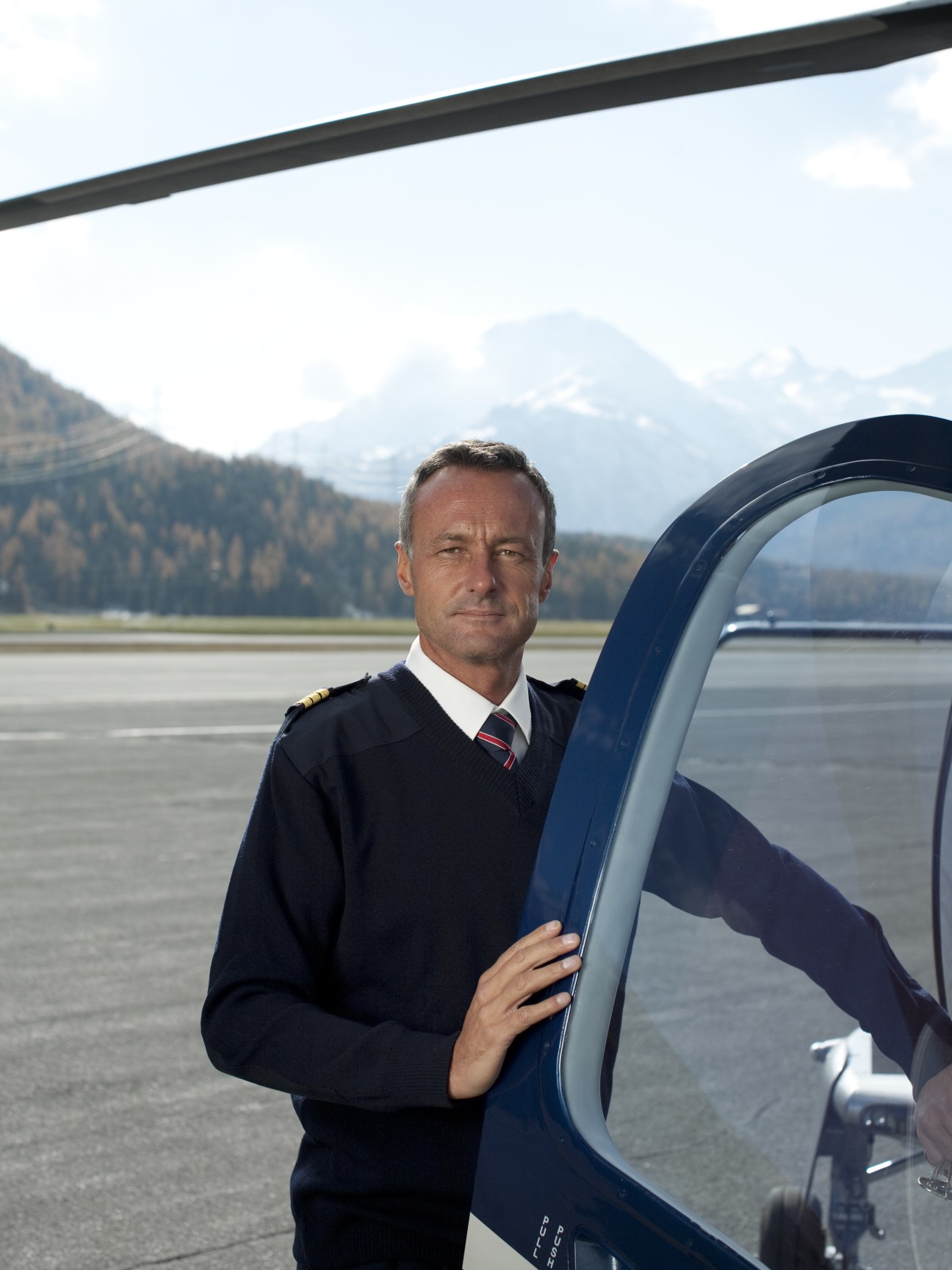 Pilot in dark uniform standing beside Swiss Jet helicopter with alpine mountains in background.