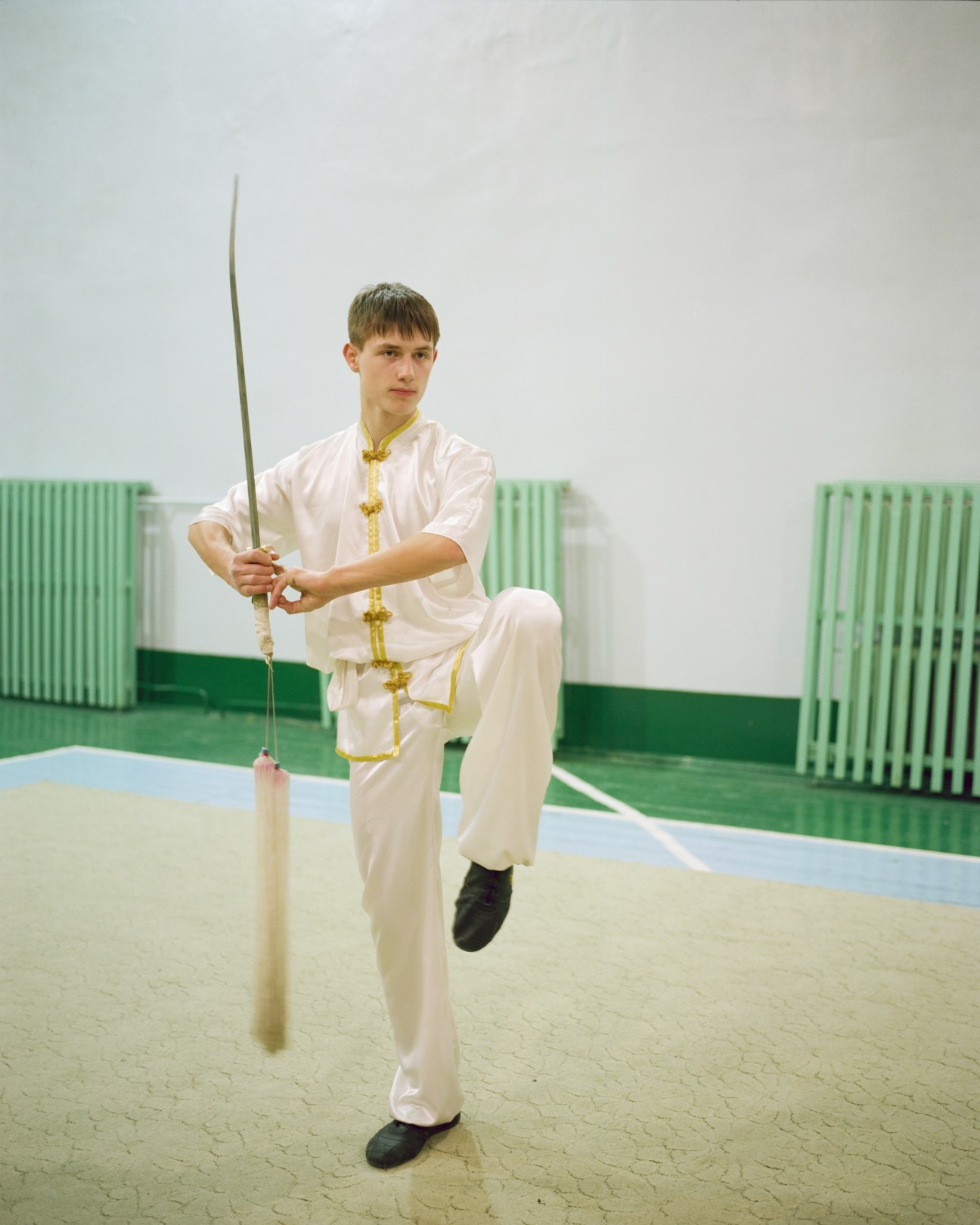 Young boy in white martial arts uniform with gold trim performs wushu with a long sword in a gymnasium.