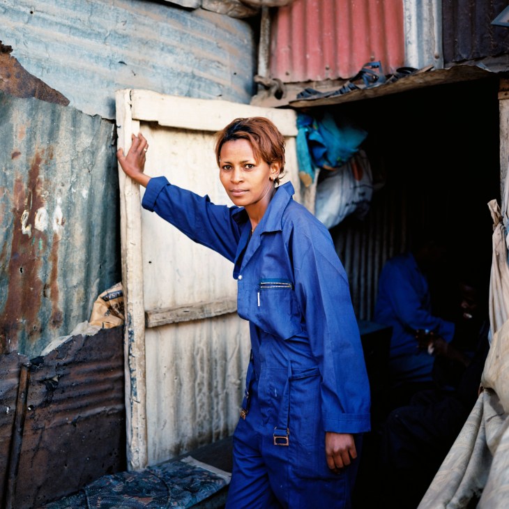 Woman in blue work clothes standing in doorway of corrugated metal structure at Medeber market.