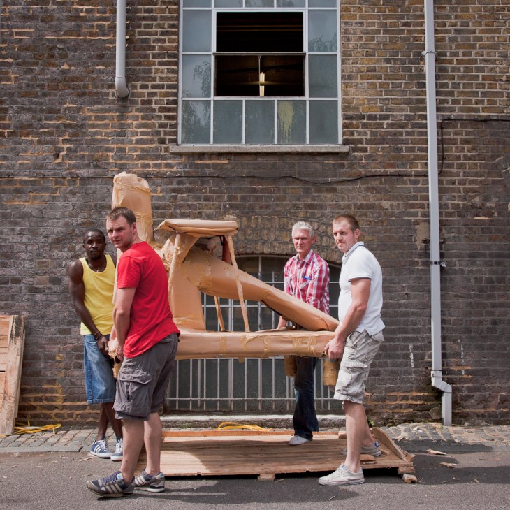 Four men carrying a large wooden sculptural artwork outside a brick building.