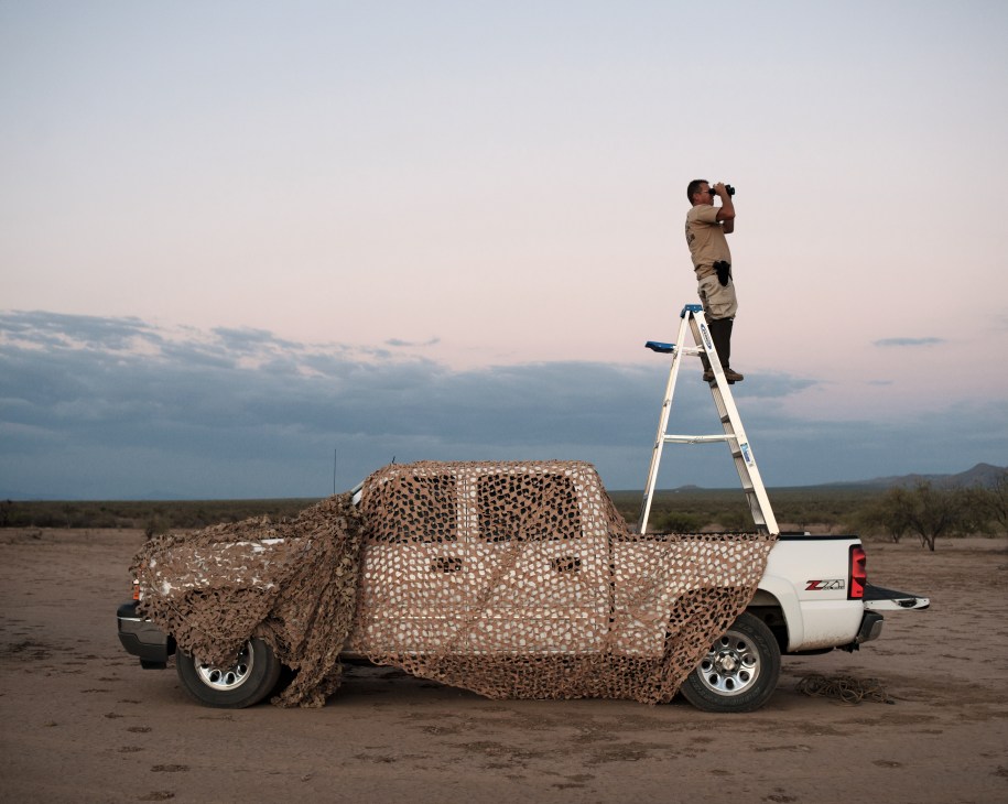 Minuteman volunteer stands on ladder atop camouflaged truck scanning desert landscape with binoculars.
