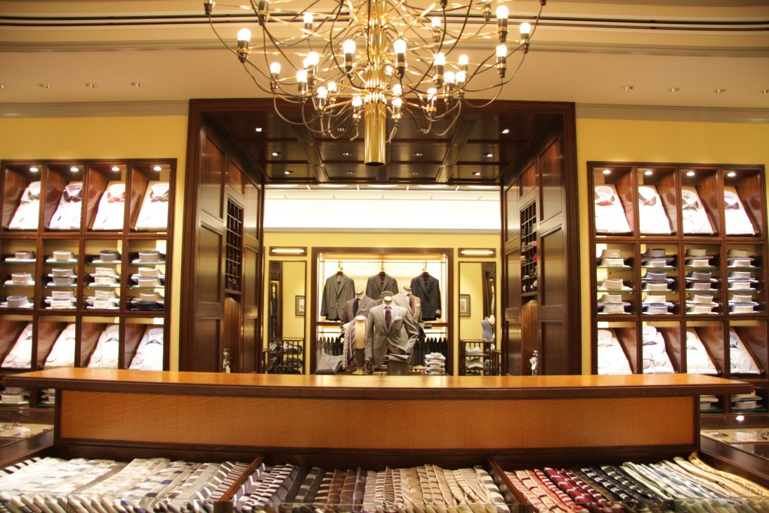 Interior of Beams House retail store with wooden display cases, brass chandelier, and menswear on mannequins.