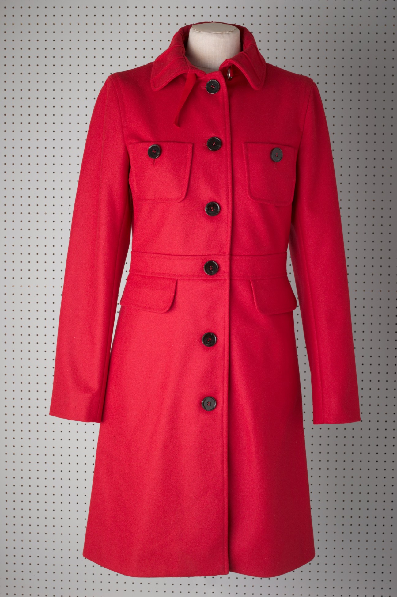 Red wool coat with black buttons and patch pockets on dress form against pegboard background.