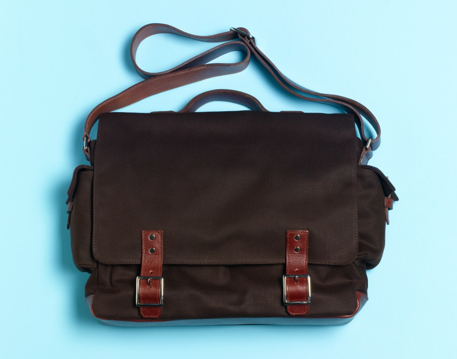 Dark brown twill messenger bag with cognac leather straps and magnetic pocket closures on light blue background.