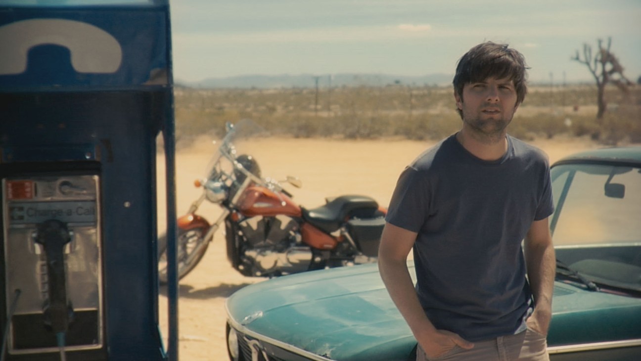 A man in a dark shirt stands at a gas station in a desert landscape with vintage cars and a motorcycle nearby.