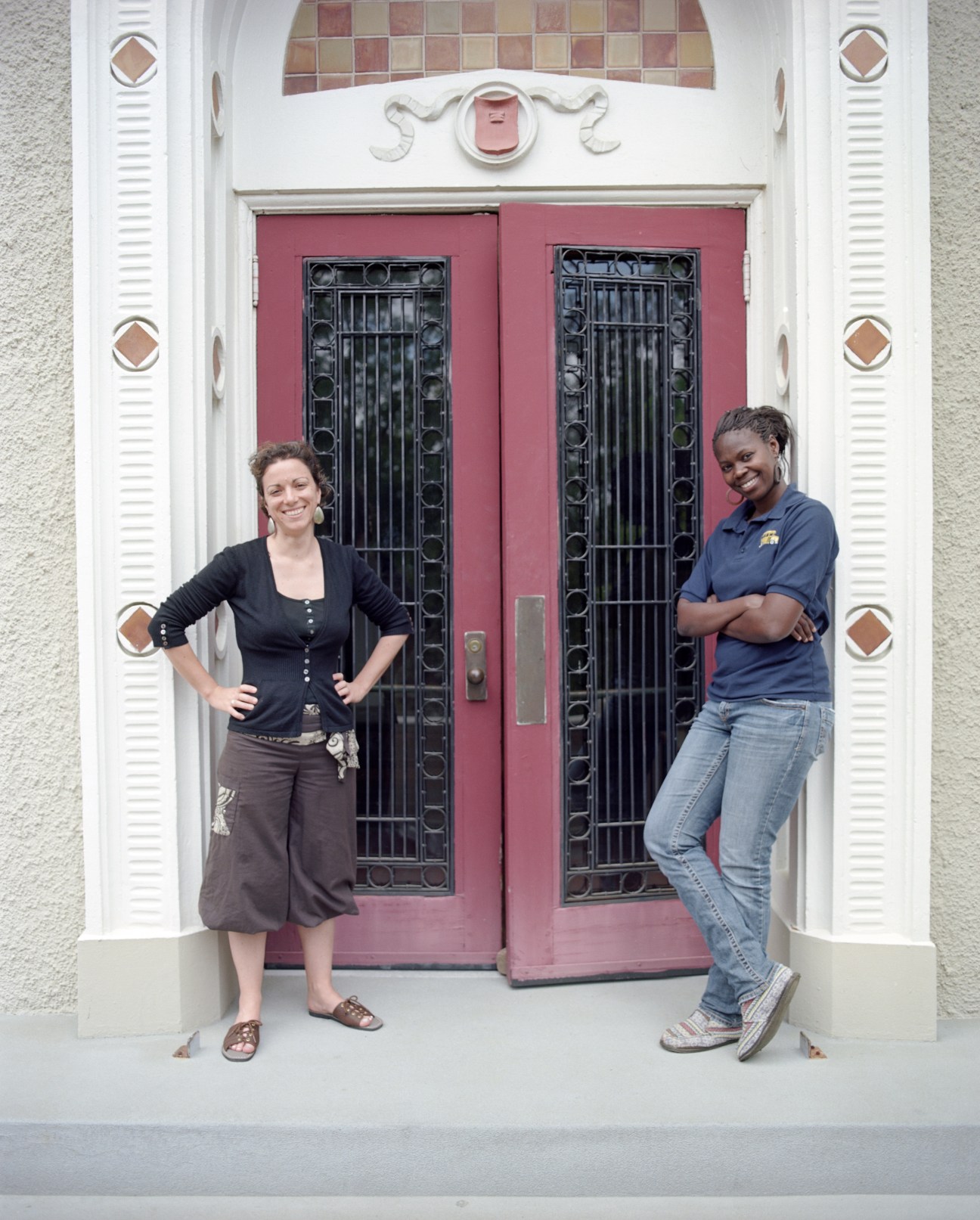 Two French teachers pose in front of a historic building with red doors and white colonial architecture.