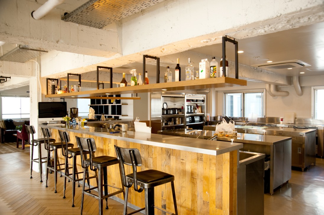 Modern shared kitchen and lounge with wooden island counter, bar seating, and open shelving displaying bottles and books.
