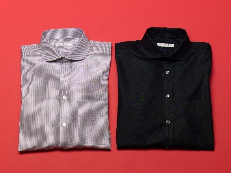 Two folded John Smedley tailored shirts on a bright red background, one light purple striped and one black.