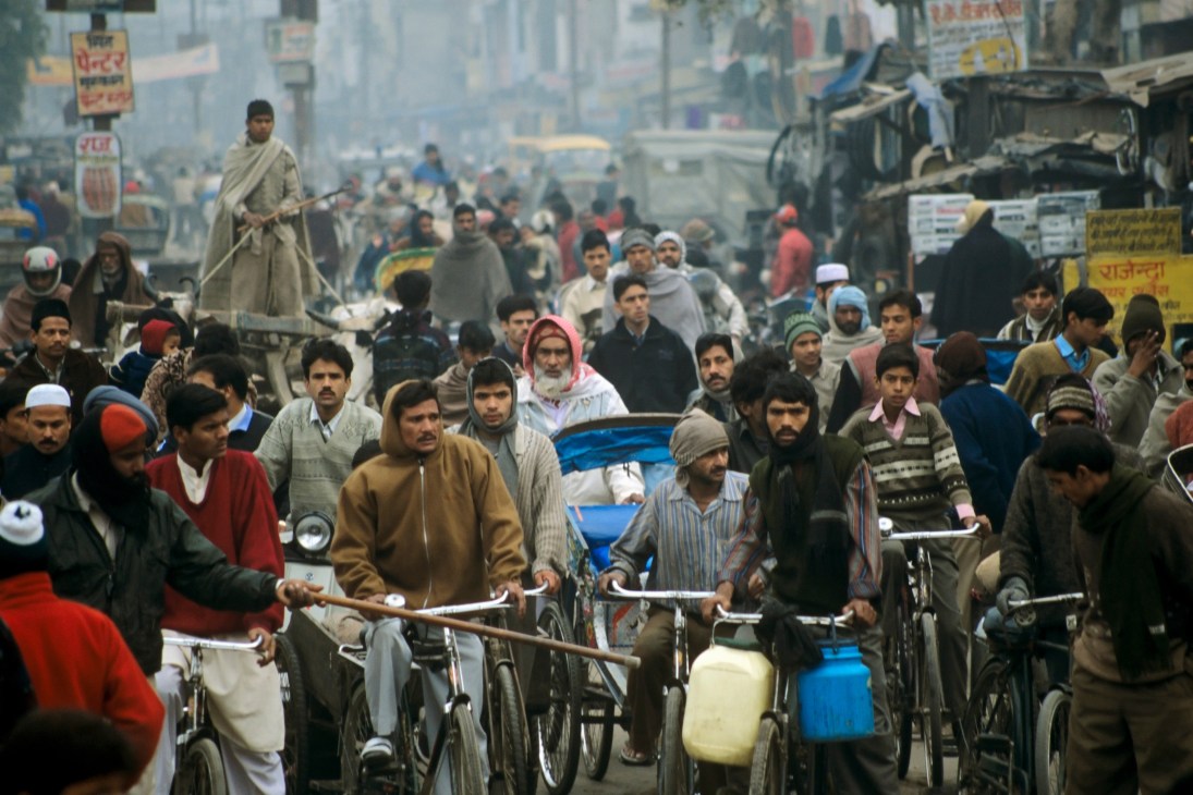 Crowded Indian street scene with pedestrians, cyclists, vehicles and Hindi signage in hazy urban environment.