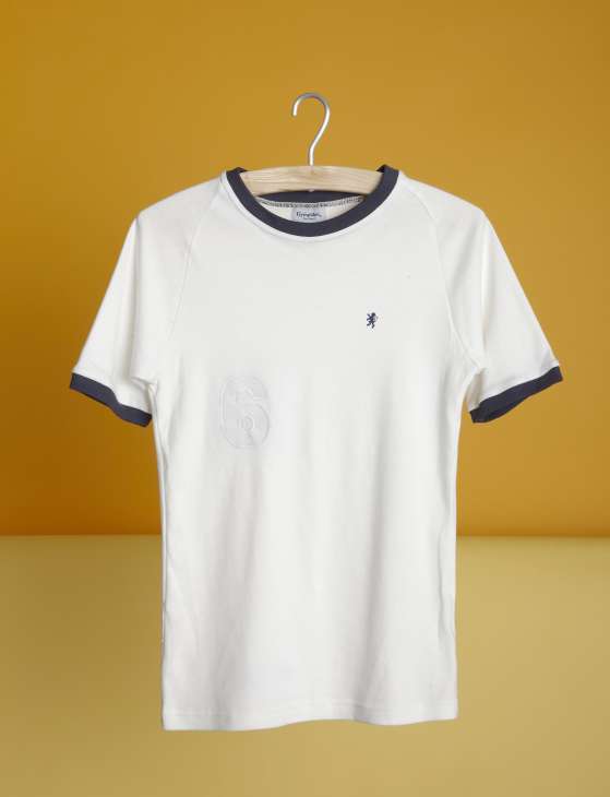 White cotton T-shirt with navy trim on neckline and sleeves, displayed on wooden hanger against mustard yellow background.