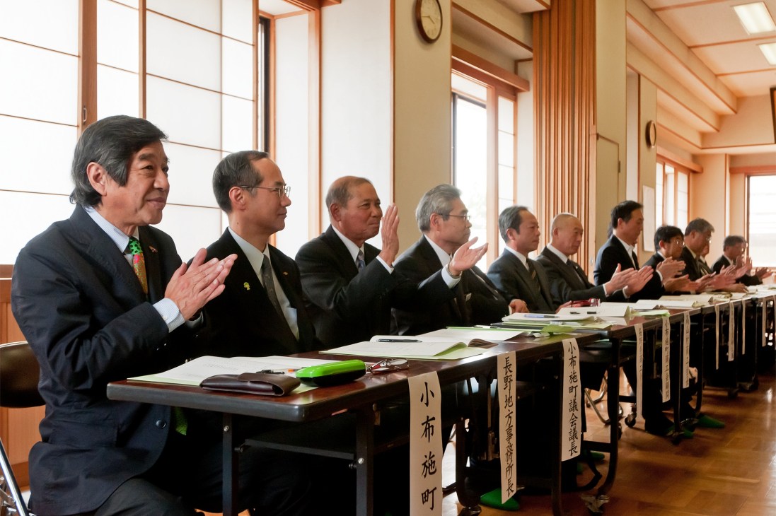Japanese officials in business attire clapping during a formal meeting in a traditional wooden hall.