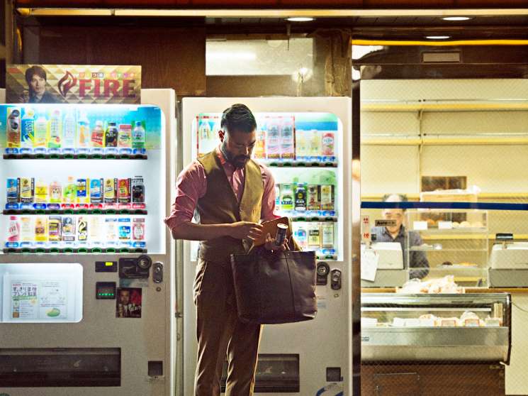 Man in designer clothing using a Japanese vending machine dispensing drinks at night on a Tokyo street.