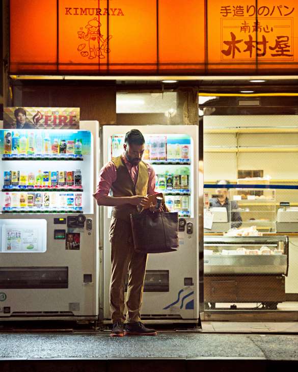 Man in designer clothing using a Japanese vending machine dispensing drinks at night on a Tokyo street.