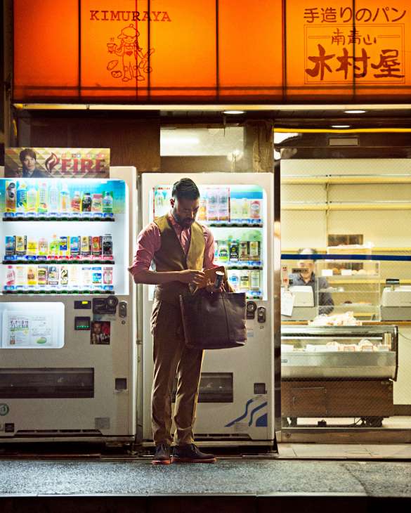 Man in designer clothing using a Japanese vending machine dispensing drinks at night on a Tokyo street.