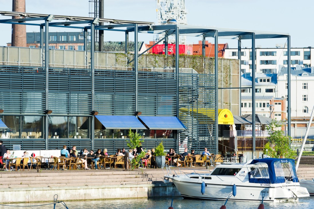 Waterfront café with blue-glass building and moored boats in Helsinki's harbor district.