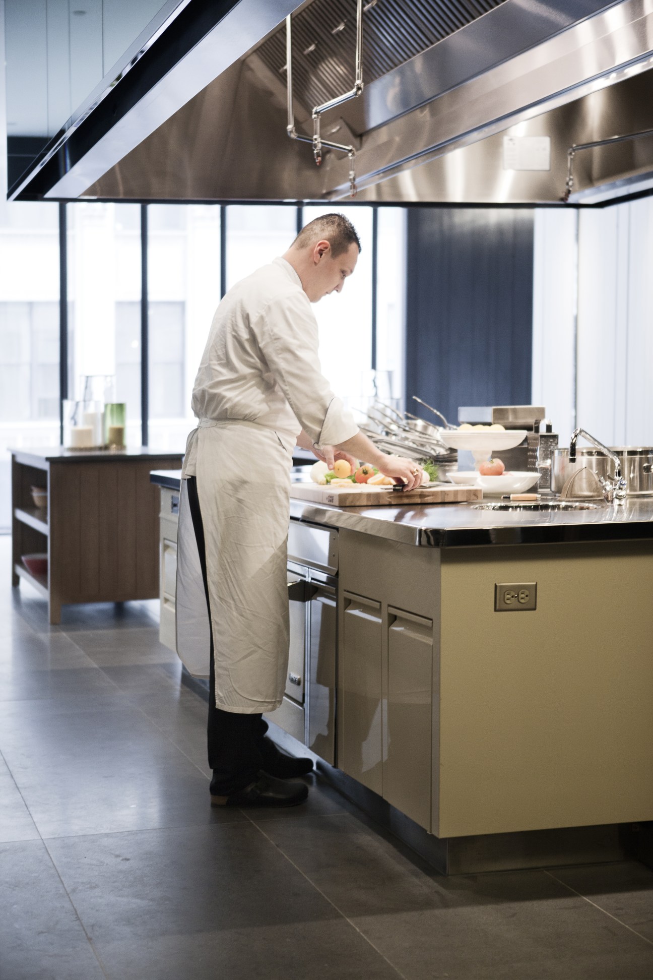 Chef in white apron preparing food at a stainless steel counter in a modern kitchen with large windows.