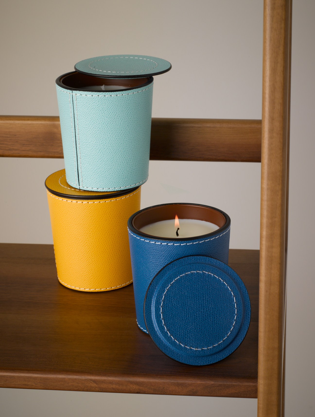 Three handmade scented candles in leather holders in blue, yellow, and light blue colors with lit wicks.