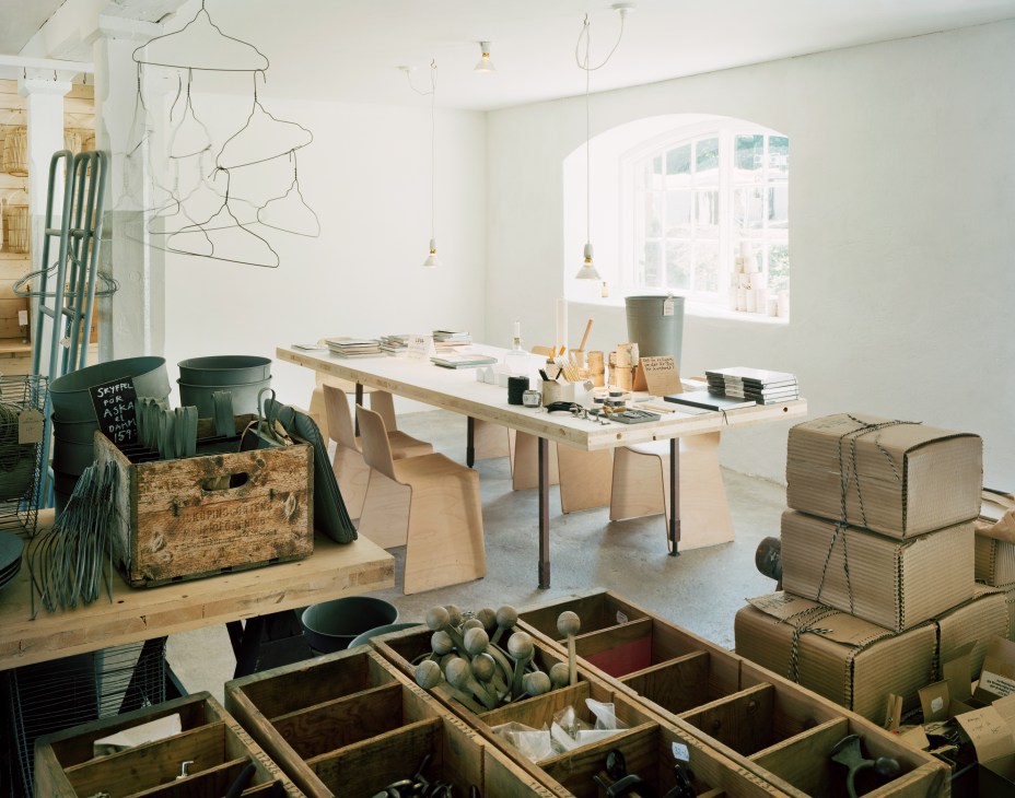 Bright studio workspace with wooden table, storage boxes, hanging wire sculptures, and gardening tools displayed in compartments.