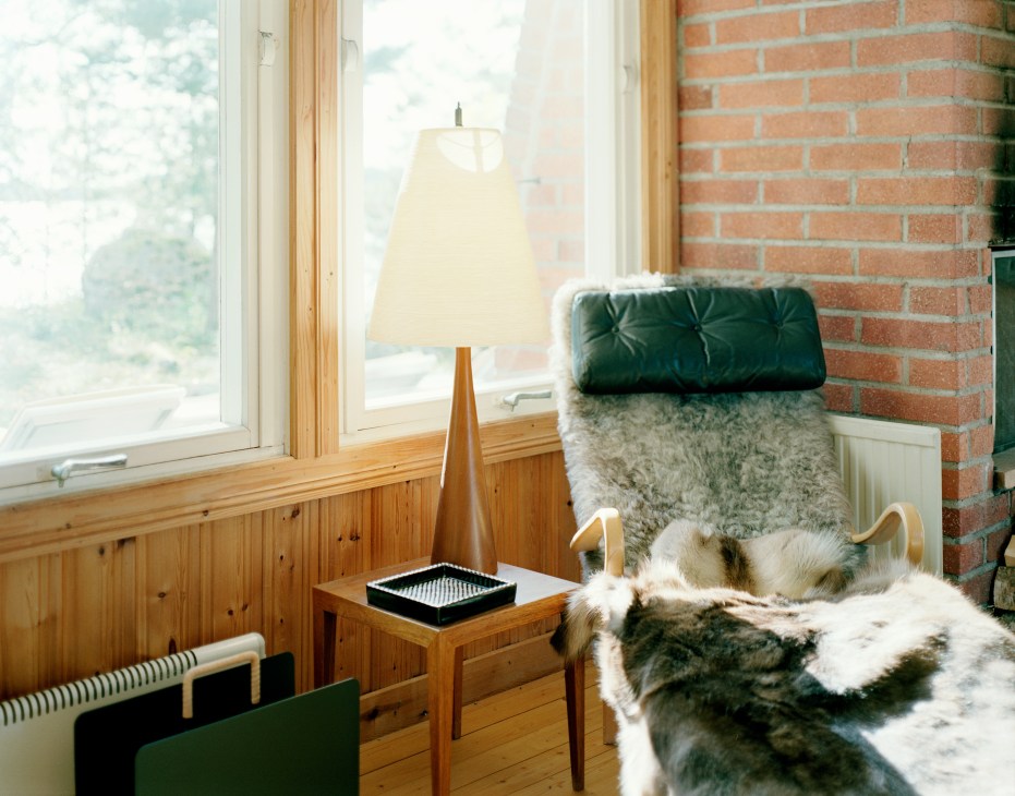 A green velvet upholstered lounge chair with furs and blankets beside a wooden side table with lamp near a window.