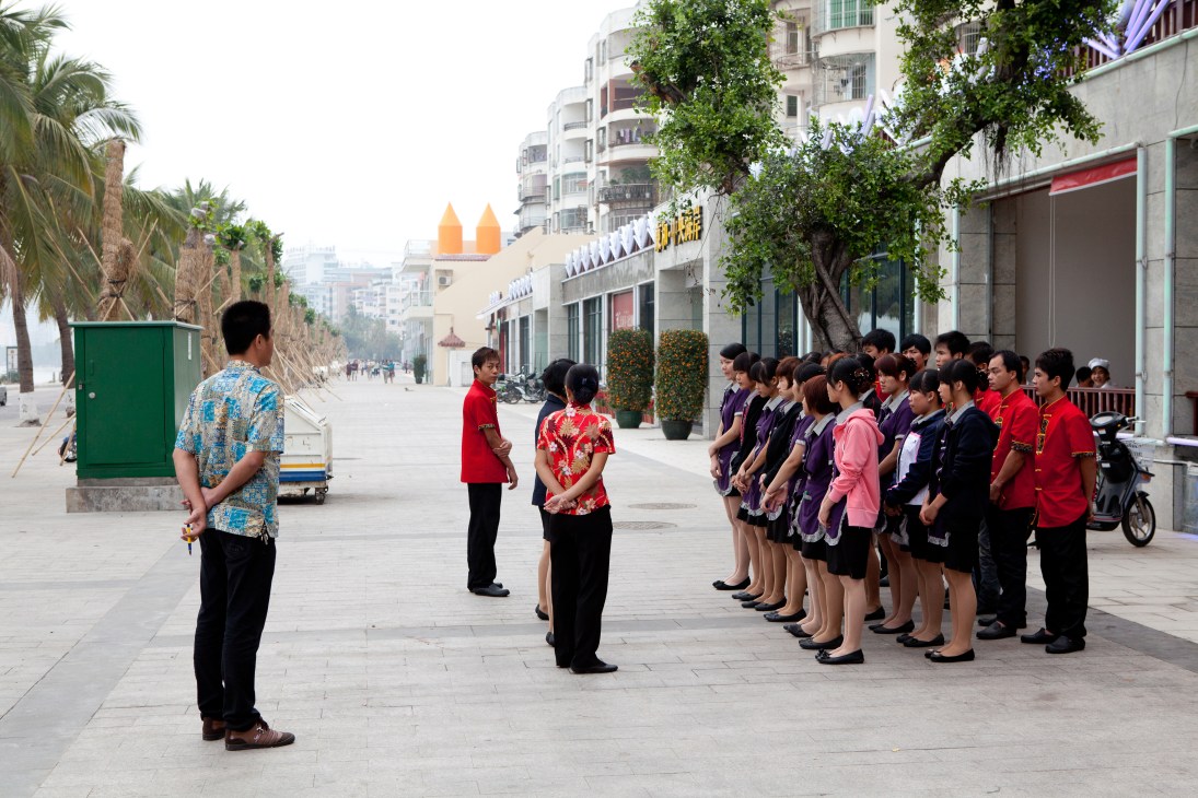 Restaurant staff in uniform standing in formation on a waterfront promenade during training in Haikou, Hainan.