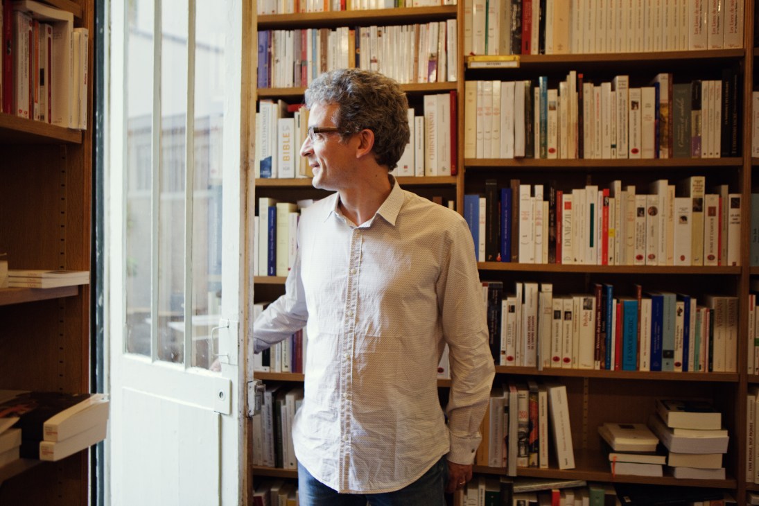 Yannick Poirier stands in Tschann Libraire bookstore surrounded by wooden shelves filled with books.