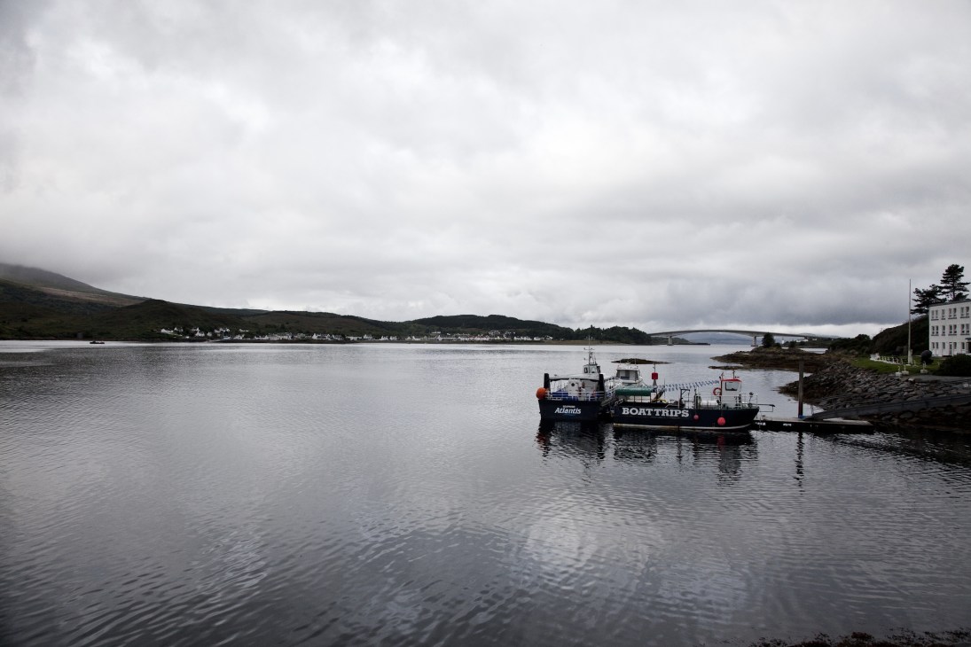 Boats moored in calm waters with a Scottish coastal village and mountains under cloudy skies.