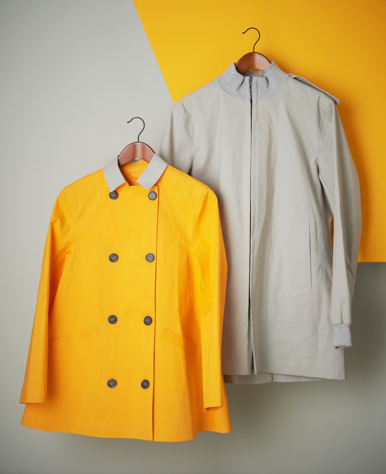Two handmade raincoats on wooden hangers against a color-blocked gray and yellow background: a bright yellow double-breasted mac and a cream-colored jacket.