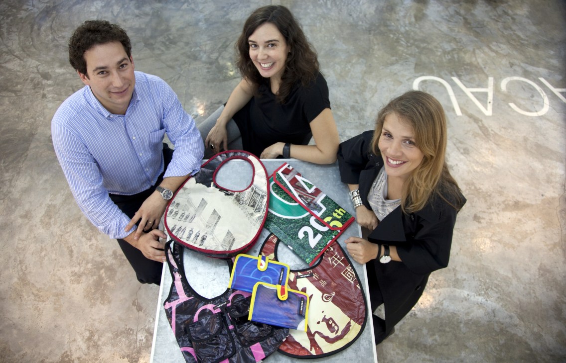 Three people display colorful bags made from recycled advertising material including circuit boards and fabric scraps.