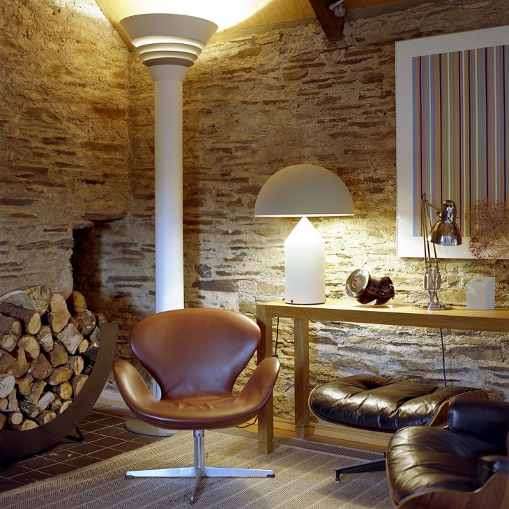 Modern living space with stone walls, brown leather chair, white dome lamp, and stacked firewood.