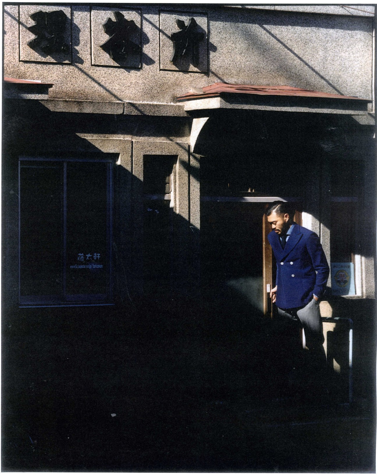 Man in blue blazer standing at entrance of traditional Tokyo barber shop Totaken with handcrafted windows above.