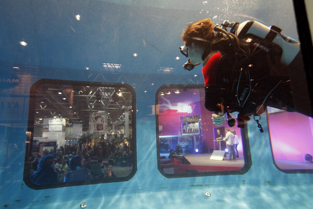 A diver in red and black diving gear with equipment inside a large transparent underwater cube at Boot Düsseldorf exhibition.