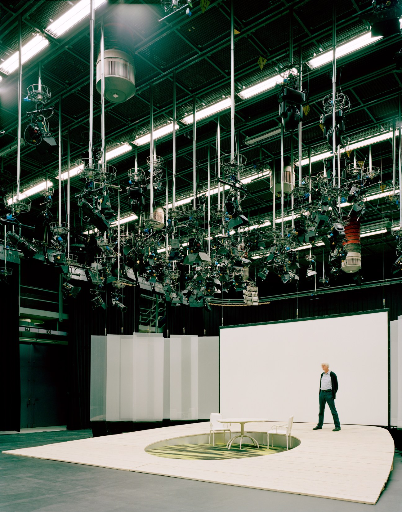 Man standing in a television studio set with green industrial ceiling framework and lighting rigs overhead.