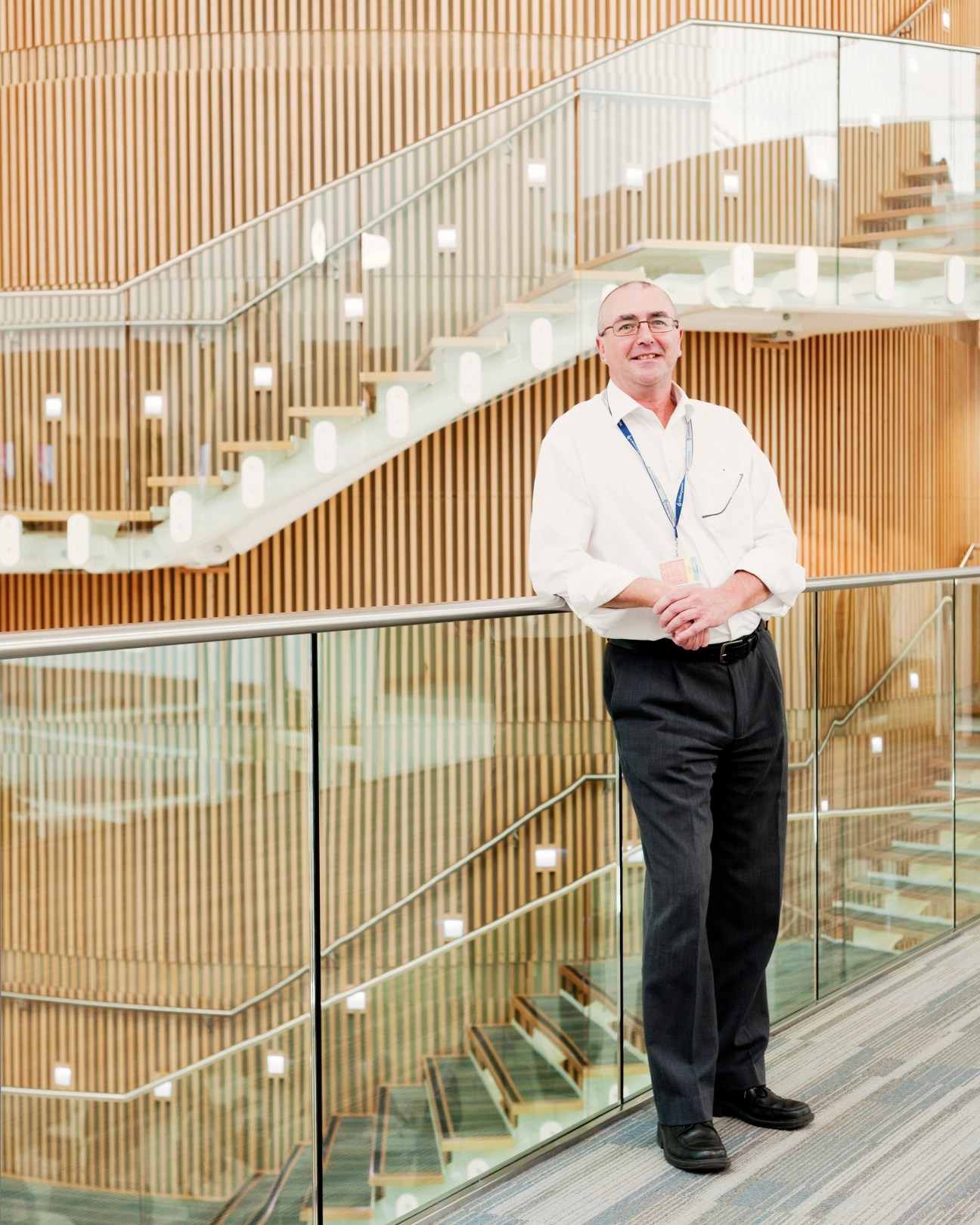 Man in white shirt and dark trousers standing at a glass railing in a modern building with wooden slat walls and stairs.